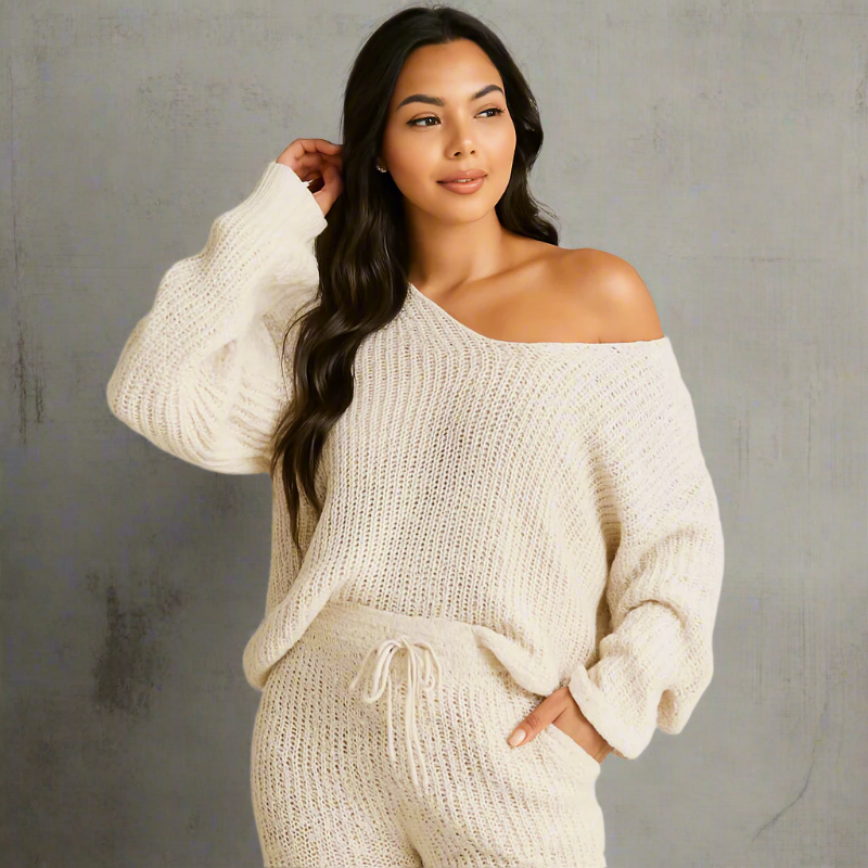 A relaxed young woman poses in an off-white chunky knit loungewear set with an oversized sweater that slips off one shoulder. Her long, wavy dark brown hair and soft pink lip add warmth to the neutral-toned setting, softly lit near sheer white curtains for a calm, cozy vibe.