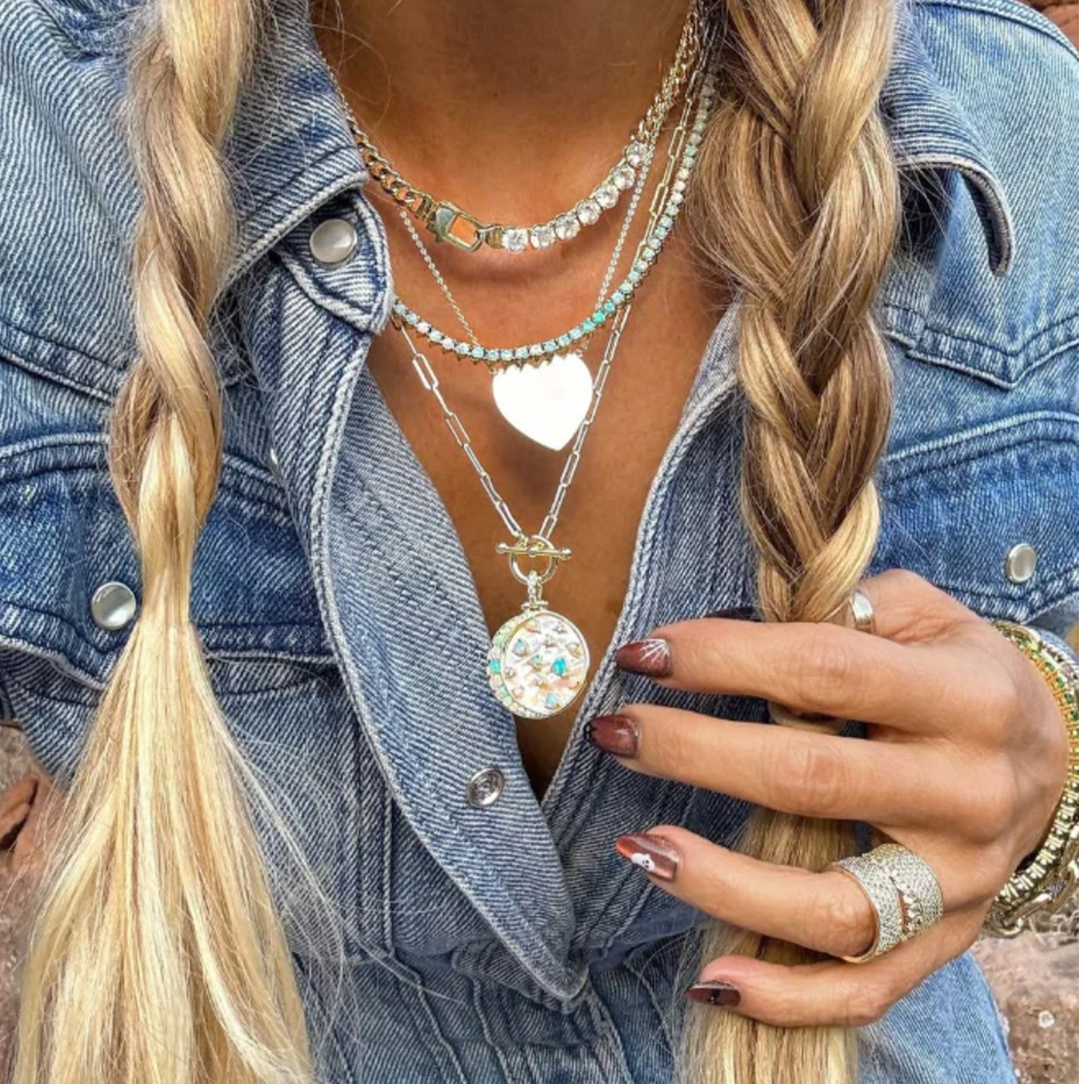 Woman wearing layered necklaces, including a heart charm and jeweled pendant, styled with a denim button-up and braided hair.