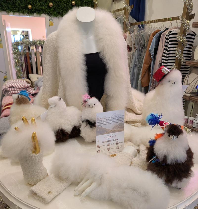 A display of alpaca-themed items, including plush toy llamas, alpaca wool mittens, a fur-trimmed shawl, and slippers, arranged on a boutique table.