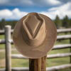 A vegan suede cowboy hat with a flat brim and arrowhead crease, displayed on a wooden fence post with a rustic countryside backdrop. Available in assorted colors to complement various styles.