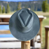 A vegan suede cowboy hat with a flat brim and arrowhead crease, displayed on a wooden fence post with a rustic countryside backdrop