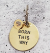 A round brass keychain engraved with "Born This Way," featuring an attached smaller charm, displayed on a textured background.