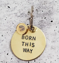 A round brass keychain engraved with "Born This Way," featuring an attached smaller charm, displayed on a textured background.