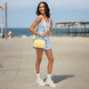 Model wearing a yellow braided woven nylon crossbody bag with an adjustable strap, standing on a boardwalk with the beach in the background.