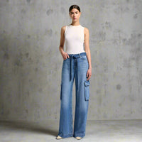Model wearing wide-leg cargo jeans in medium blue denim with front cargo pockets and a matching denim tie belt, paired with a fitted white sleeveless top and cream-colored heels.