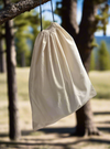 A cotton muslin drawstring dust bag hanging from a tree branch, designed for hat protection and storage.