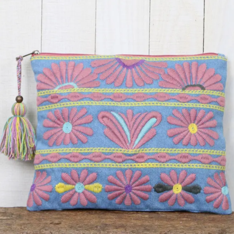 Denim clutch bag with pink and pastel floral embroidery, yellow stitching, and multicolored tassel zipper pull on a rustic wood surface.

