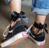 Close-up of black fishnet ankle socks with satin bow detail worn with black Vans sneakers and cuffed jeans.

