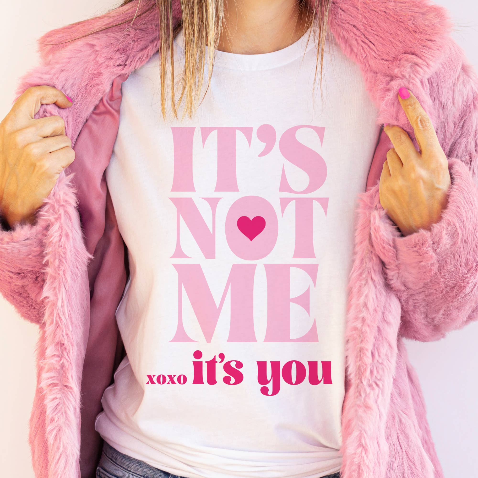A white graphic t-shirt with bold pink lettering reading "It’s Not Me, It’s You" featuring a heart detail, styled with a pink faux fur coat.