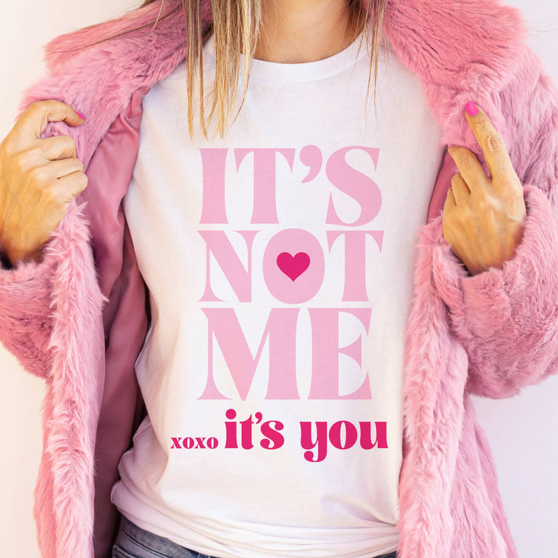 A white graphic t-shirt with bold pink lettering reading "It’s Not Me, It’s You" featuring a heart detail, styled with a pink faux fur coat.
