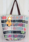 Colorful patchwork denim bucket bag with neon embroidery, vegan leather handles, and tassel charm hanging against white wood panel background.