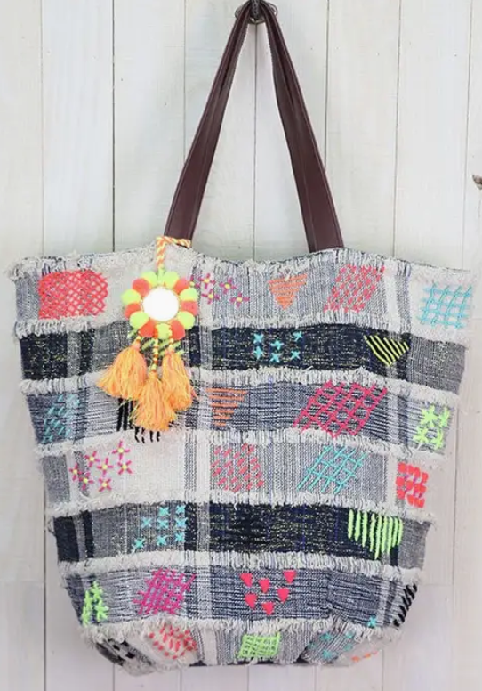 Colorful patchwork denim bucket bag with neon embroidery, vegan leather handles, and tassel charm hanging against white wood panel background.
