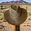 A dusty olive-green vegan suede cowboy hat with a flat brim and Western crown, resting on a wooden fence post in a rugged desert landscape with mountains in the background.