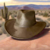 A dusty olive-green vegan suede cowboy hat with a flat brim and Western crown, resting on a boulder in a rugged desert landscape with mountains in the background.