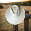 A creme vegan suede rancher hat with a wide flat brim, perched on a wooden fence post at sunset with a scenic countryside backdrop.