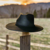 A black vegan suede rancher hat with a wide flat brim, perched on a wooden fence post at sunset with a scenic countryside backdrop.