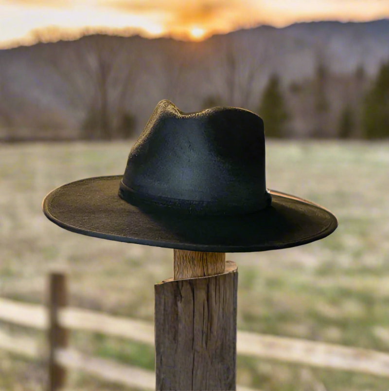 A black vegan suede rancher hat with a wide flat brim, perched on a wooden fence post at sunset with a scenic countryside backdrop.