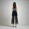 Back view of black high-rise jeans with sheer lace paneling from thigh to hem, styled with a grey tank top and heels