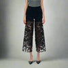 Full front view of black high-rise jeans with rhinestone embellishments and sheer lace cutout legs, styled with pointed black heels and a grey top