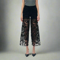 Full front view of black high-rise jeans with rhinestone embellishments and sheer lace cutout legs, styled with pointed black heels and a grey top