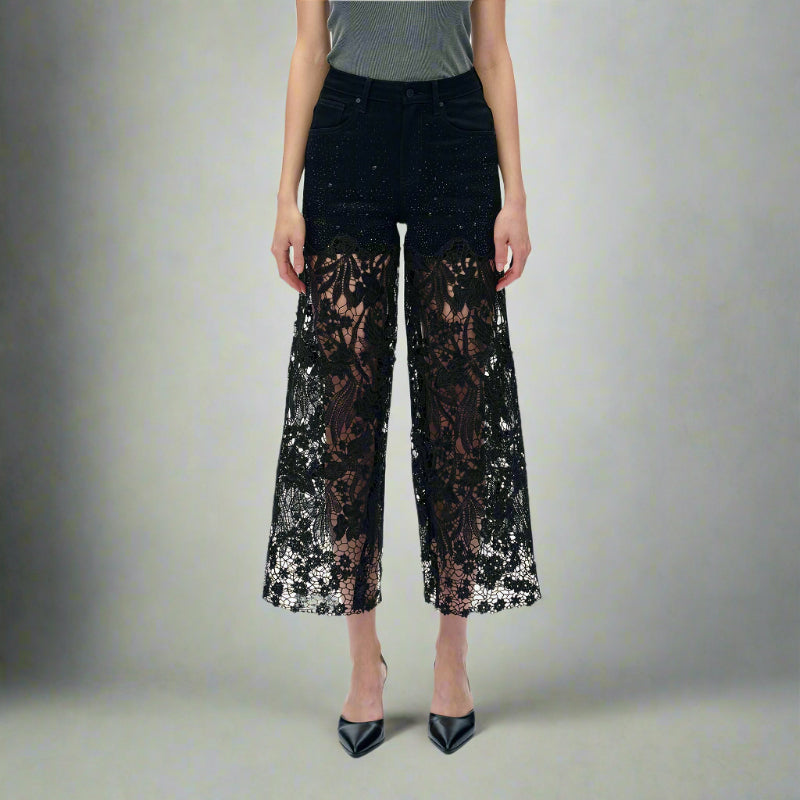 Full front view of black high-rise jeans with rhinestone embellishments and sheer lace cutout legs, styled with pointed black heels and a grey top
