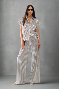 Woman wearing a metallic silver pleated short-sleeve button-up shirt styled with matching pants, white belt, and sunglasses