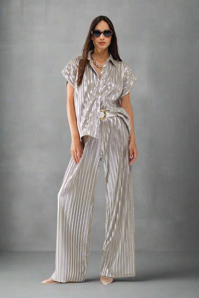 Woman wearing a metallic silver pleated short-sleeve button-up shirt styled with matching pants, white belt, and sunglasses