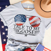 Light blue graphic t-shirt featuring aviator sunglasses with an American flag design and "Talk to Me Goose" text in bold, vintage-style print.