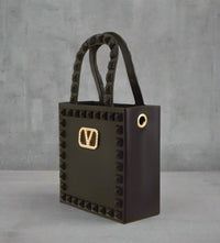 Mini black jelly handbag with studded edges and a gold-tone “V” buckle on the front, featuring textured top handles and gold grommet detail.