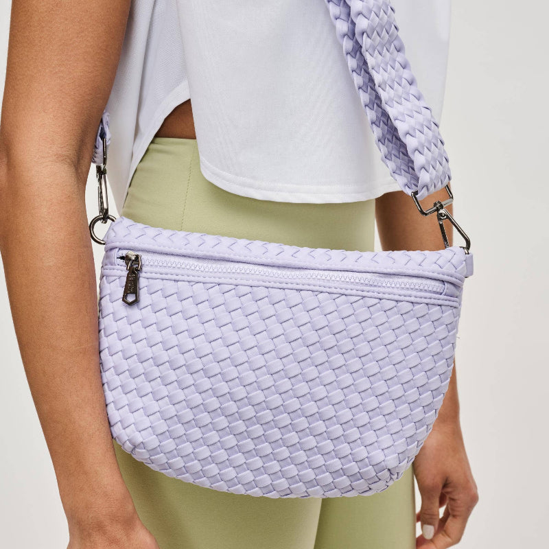 Model wearing a woven neoprene crossbody bag in a soft pastel shade, featuring an adjustable strap, zipper closure, and modern gunmetal hardware.