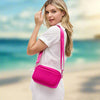 Model wearing a hot pink woven neoprene crossbody bag with an adjustable strap, standing in front of a beachside backdrop.