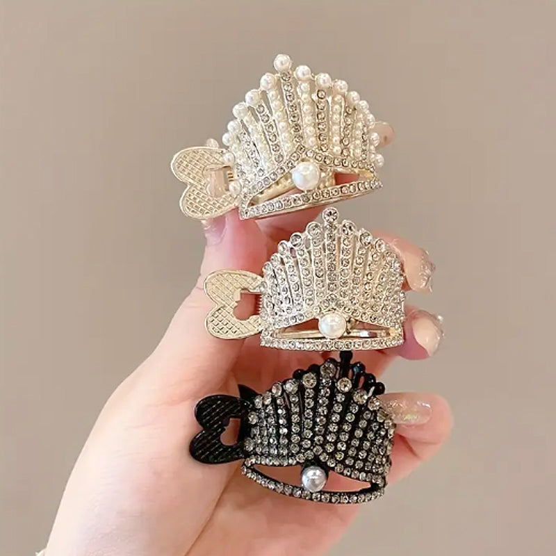 Hand holding three rhinestone high ponytail clips in gold, silver, and black, featuring a tiara-inspired design with crystal embellishments and pearl accents.